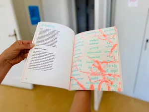 Hand hält ein aufgeschlagenes Buch mit links gedrucktem Fließtext und rechts einer Seite mit überlappenden, in verschiedenen Grüntönen gedruckten Wörtern und roten, abstrakten Linien.