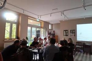 Gruppe von Menschen sitzt an einem langen Tisch in einem hellen Raum mit großen Fenstern und einer Projektionsfläche an der Wand