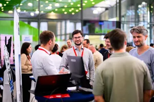 Teilnehmende tauschen sich an einem Messestand des 2. Brandenburger Digitaltags aus. Im Vordergrund steht eine Person mit rotem Schlüsselband hinter einem technischen Ausstellungsobjekt.