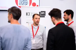 Ein Referierender mit rotem Schlüsselband spricht vor einem Banner des KI-Service-Zentrums des Hasso-Plattner-Instituts, im Raum mehrere Teilnehmende.