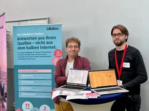 Das InNoWest KI-Chatbot-Team präsentiert sich auf dem Markt der Möglichkeiten. Zwei Teammitglieder stehen lächelnd hinter einem Stehtisch mit zwei Laptops. Links daneben steht das Roll-up-Banner.