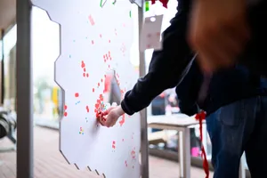 Eine weiße Deutschlandkarte auf einem mobilen Gestell vor einer Backsteinwand, bestückt mit zahlreichen roten, blauen, grünen und gelben Punkten.