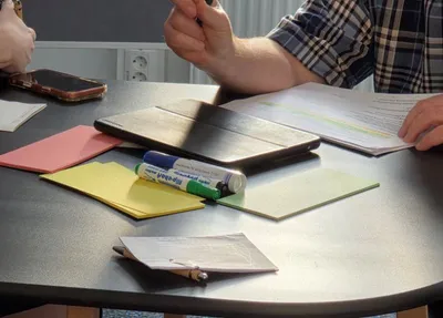 Tisch mit Tablet, mehreren bunten Notizzetteln, zwei Stiften, einem Smartphone und zwei Personen, von denen eine ein Dokument hält und mit der Hand gestikuliert