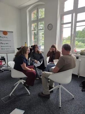 Vier Personen sitzen in einem hellen Raum in einem Stuhlkreis und unterhalten sich, im Hintergrund ein Flipchart mit handschriftlichen Notizen und große Fenster mit Blick ins Grüne