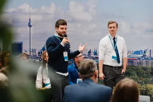 Ein Mann im dunklen Pullover spricht gestikulierend ins Mikrofon, ein zweiter Mann im weißen Hemd steht daneben; im Hintergrund eine Berlin-Fotografie, am Rand sitzende Personen.