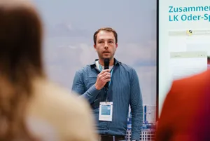  Ein Mann mit Namensschild und Mikrofon spricht vor einer Leinwand; rechts ist ein Folienausschnitt mit Text und einer digitalen Anwendung zu sehen.