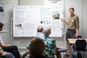 Zu sehen ist eine Mann vor einem Poster, die etwas dazu erzählt