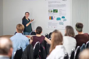 Zu sehen ist eine Frau vor einem Poster, die etwas dazu erzählt