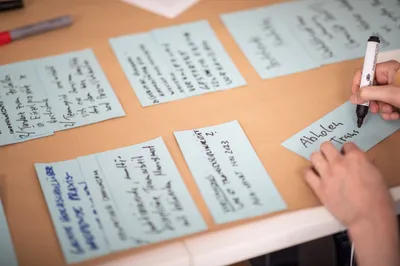 Das Foto zeigt eine Workshop Situation bei der unterschiedliche Statements auf einem Papier liegen. Ein Statement wird gerade auf eine Karte geschrieben. 