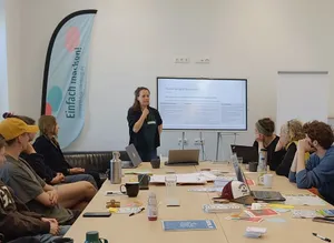 Eine Gruppe von Menschen sitzt um einen Tisch, während eine Frau eine Präsentation hält.  