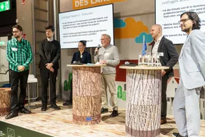 5 Personen stehen auf einer Bühne mit runden Stelltischen aus Holz und vor 2 Bildschirmen