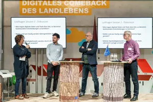 4 Personen stehen auf einer Messebühne vor 2 Bildschirmen und 2 Holztischen