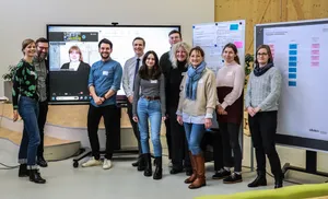 Gruppenbild mit elf Personen, die vor Stellwänden und einem großen Bildschirm stehen. Eine Person ist digital zugeschaltet