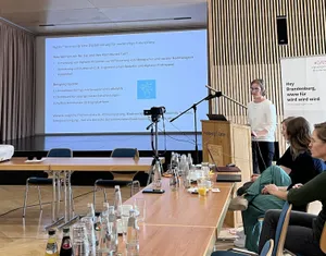Man sieht eine Frau am Stehpult sprechen vor einem Publikum und einer großen Leinwand