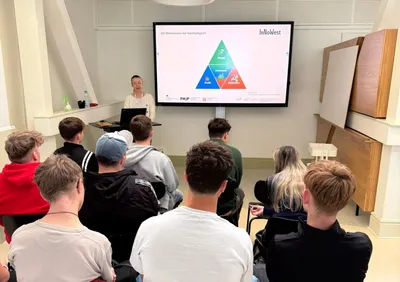 Zu sehen ist eine Gruppe von Schüler*innen, die vor einer Vortragenden Person sitzen mit einem großen Smartboard an der Wand