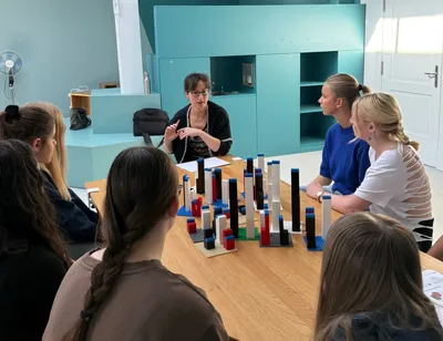 Zu sehen sind Schüler*innen an einem Tisch mit Bausteinen, die einer Frau beim redden zuhören