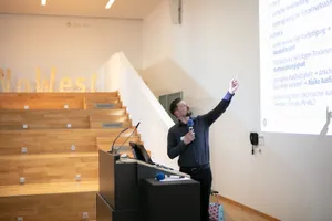 Ein Mann steht an einem Rednerpult und ist seinen Präsentationsfolien zugewand und zeigt darauf.