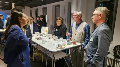 Zu sehen ist eine Gruppe von Menschen, die um einen Tisch herum stehen und sich unterhalten