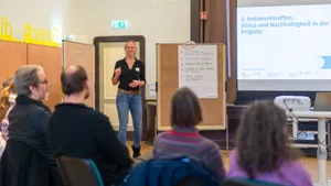 Zu sehen ist eine Frau, die vor Menschen steht, die auf Stühlen sitzen. Rechts wird eine Präsentation mit einem Beamer eingeblendet.