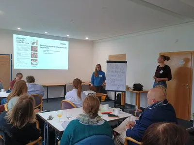 Zu sehen ist ein Raum mit Menschen, die zu einem Flipchart und einer an die Wand gestrahlte Power Point Präsentation schauen. 2 Frauen stehen neben dem Flipchart und moderieren.