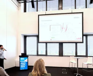 Person hält Mikrofon und präsentiert vor Leinwand mit Diagramm und Text 'daten kombinieren' in hellem Raum mit großen Fenstern.