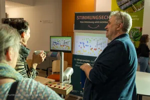 Mehrere Personen stehen vor einem Bildschirm und einem Banner mit der Überschrift STIMMUNGSECHO in einem Innenraum mit orangefarbener Wand.