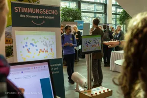 Mehrere Personen stehen in einem hellen Raum mit großen Fenstern, im Vordergrund ein Bildschirm mit farbigen Punkten und ein Banner mit der Überschrift 'STIMMUNGSECHO'.
