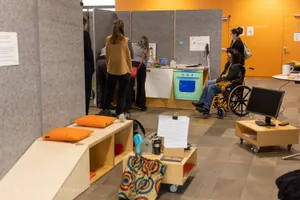 Mehrere Personen stehen vor einer Stellwand mit einem Bild eines blauen Ofens, eine Person sitzt im Rollstuhl, im Vordergrund ein niedriger Tisch mit Kissen und einem Monitor auf Rollen.