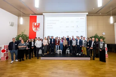 Große Gruppe von Menschen steht auf einer Bühne vor einer Leinwand mit Logos und Text, links hängt ein rotes Banner mit weißem Wappen.