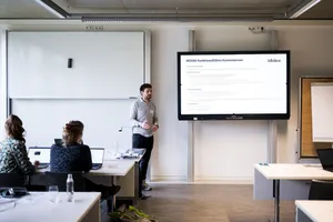 Person steht vor großem Bildschirm mit Präsentation, zwei Personen sitzen an Tischen und schauen zu.