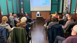 Mehrere Personen sitzen in einem Raum mit grünen Wänden und Holzfußboden und schauen auf eine Leinwand mit Präsentation.