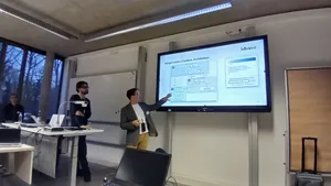 Person zeigt auf eine Präsentationsfolie mit Diagramm in einem modernen Seminarraum, weitere Personen hören zu.