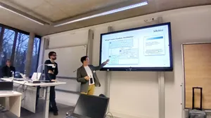 Person zeigt auf eine Präsentationsfolie mit Diagramm in einem modernen Seminarraum, weitere Personen hören zu.