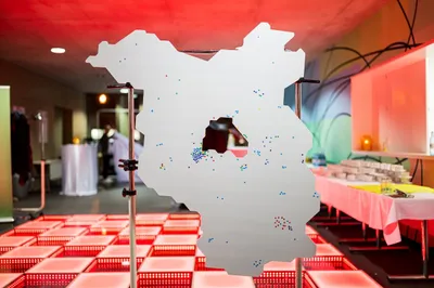 Große, weiße Karte mit vielen kleinen farbigen Punkten, montiert auf Ständern in einem Raum mit roter Beleuchtung und Tischen im Hintergrund.