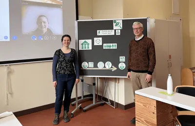 Zwei Personen stehen vor einer Tafel mit mehreren Piktogrammen und Pfeilen, im Hintergrund ist eine Projektion mit einer weiteren Person zu sehen.