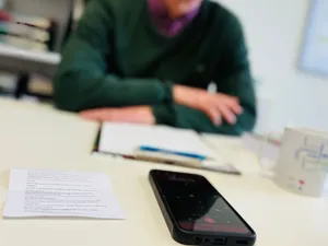 Ein Mensch sitzt an einem Tisch auf dem ein Handy eine Sprachaufzeichnung macht.