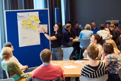 Frau steht vor Pinnwand mit Notizzetteln und zeigt darauf, mehrere Personen sitzen an Tischen und hören zu