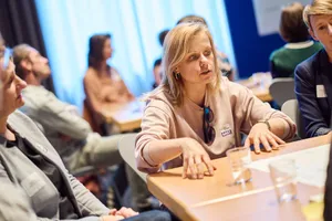 Frau mit hellbraunen Haaren und rosa Pullover sitzt an einem Tisch und gestikuliert mit den Händen