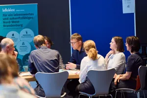 Gruppe von Menschen sitzt an einem Tisch und diskutiert in einem Raum mit blauem Trennwand und einem Plakat mit Text zu Nord-West-Brandenburg