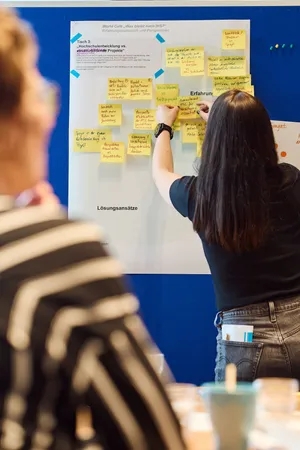 Frau mit dunklen Haaren heftet gelbe Notizzettel an eine weiße Pinnwand vor blauem Hintergrund, im Vordergrund unscharfe Person mit gestreiftem Oberteil