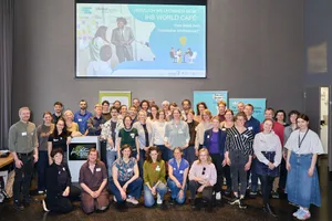 Gruppenfoto von etwa 40 Personen vor einer Leinwand mit der Projektion 'HERZLICH WILLKOMMEN BEIM IHS WORLD CAFÉ' in einem Veranstaltungsraum