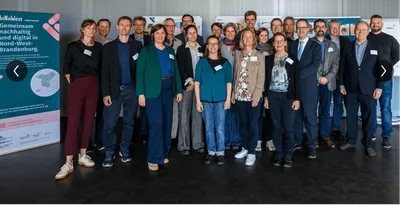 Gruppe von Menschen steht in einem Raum vor einer Wand mit einem Banner links im Bild