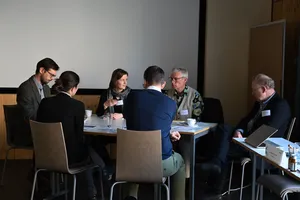 Sechs Personen sitzen an einem Tisch in einem Besprechungsraum, einige mit Getränken und Unterlagen vor sich