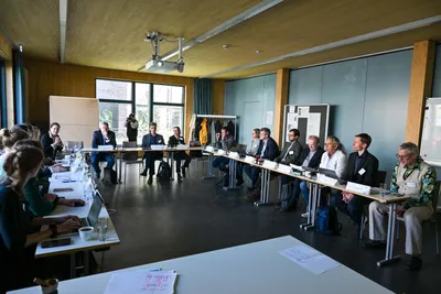 Personen sitzen an zwei gegenüberstehenden langen Tischen in einem hellen Raum mit Holzdecke und großen Fenstern