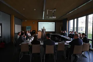 Person steht vor einem Konferenztisch, an dem mehrere Personen sitzen, im Raum mit großen Fenstern und einer Präsentation auf einer Leinwand