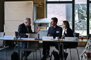 Drei Personen sitzen an einem Tisch mit Namensschildern und unterhalten sich in einem hellen Raum mit großen Fenstern.