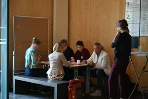 Gruppe von fünf Personen sitzt und steht um einen Tisch in einem Raum mit Holzvertäfelung, eine Person nutzt ein Smartphone