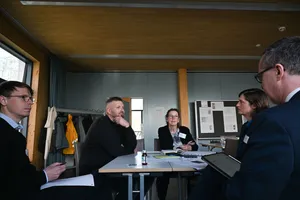 Fünf Personen sitzen an einem Tisch in einem Besprechungsraum, einige mit Notizblöcken und Getränken, im Hintergrund Garderobe und Pinnwand.