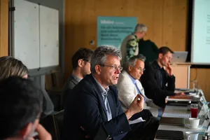 Mehrere Personen sitzen an einem langen Tisch in einem Besprechungsraum, eine Person im Vordergrund gestikuliert mit der Hand
