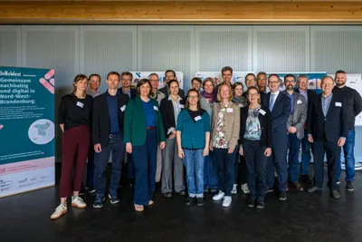 Gruppe von Menschen steht in einem Raum vor einer Wand mit vertikalen Lamellen und einem großen Informationsplakat links